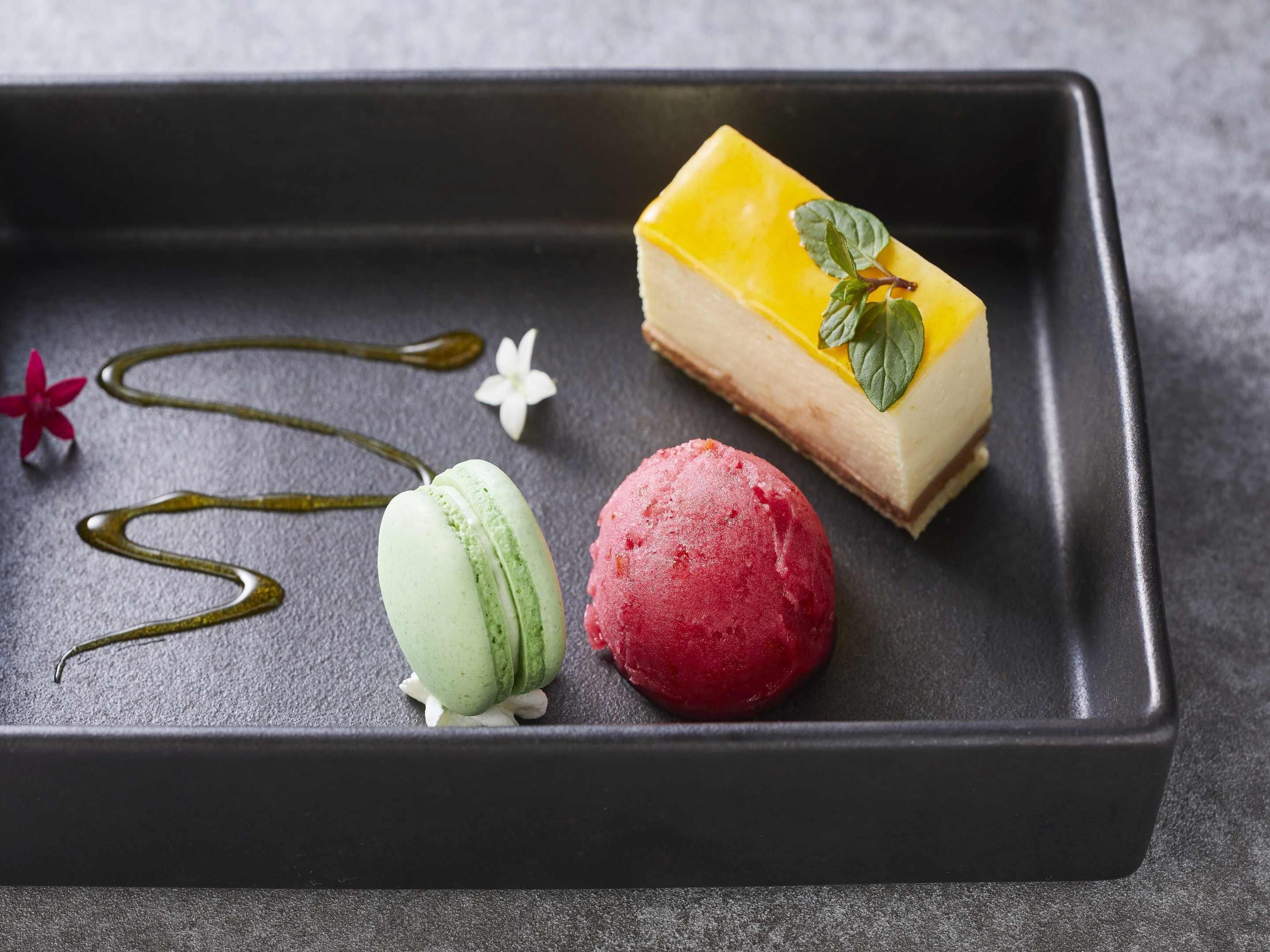 A dessert plate with a yellow cake, green and red macarons, and a drizzle of sauce.