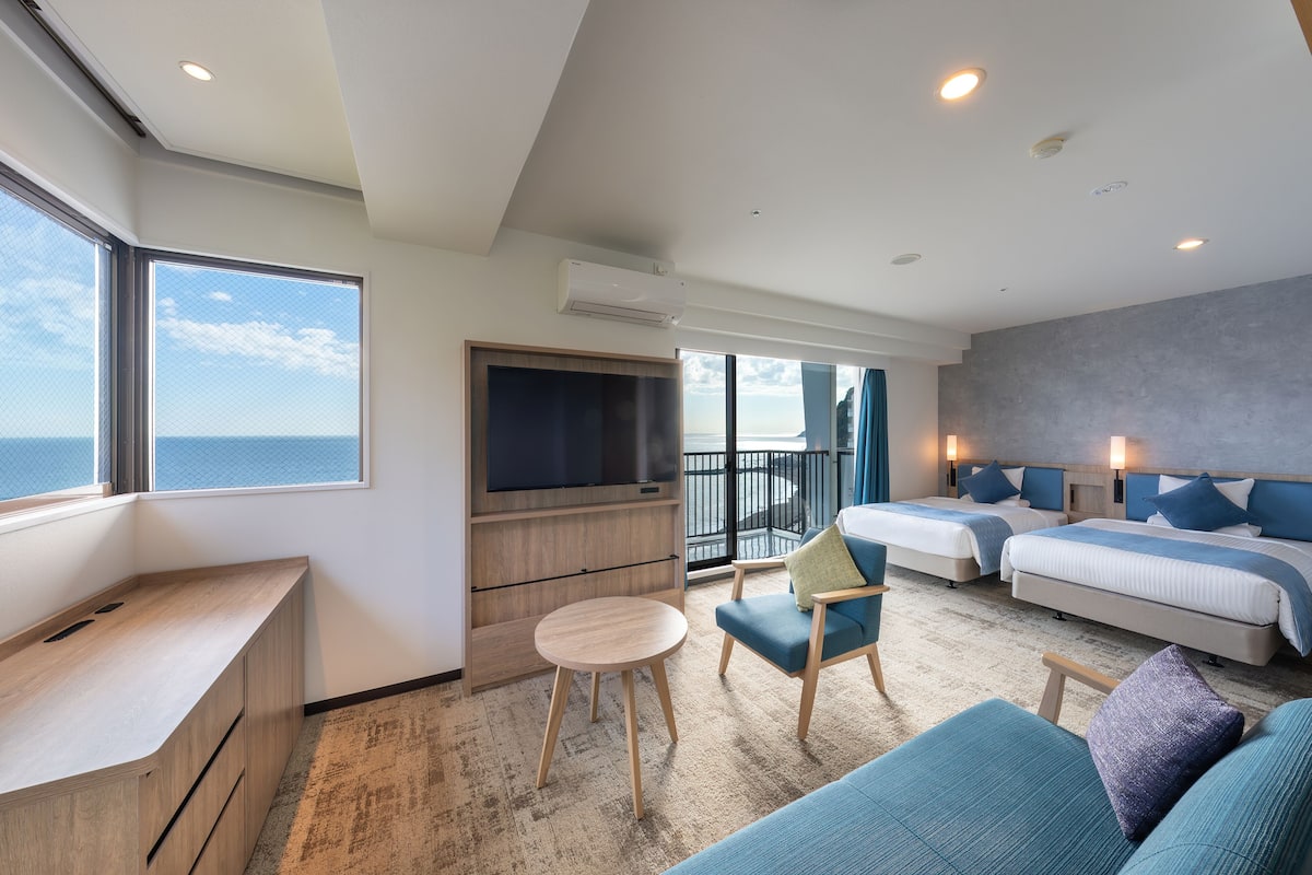 Una habitación de hotel moderna con un ventanal grande, un escritorio, dos camas y un balcón.