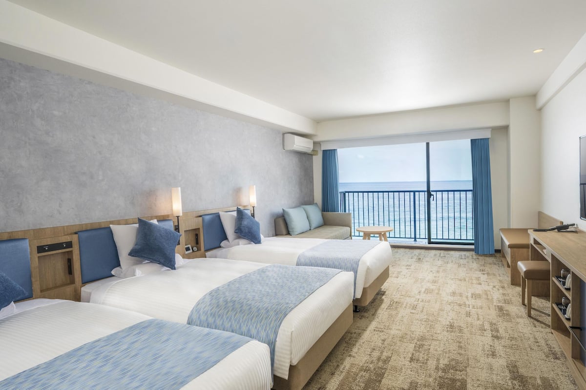 Habitación de hotel con dos camas, un escritorio y un balcón con vista al mar.