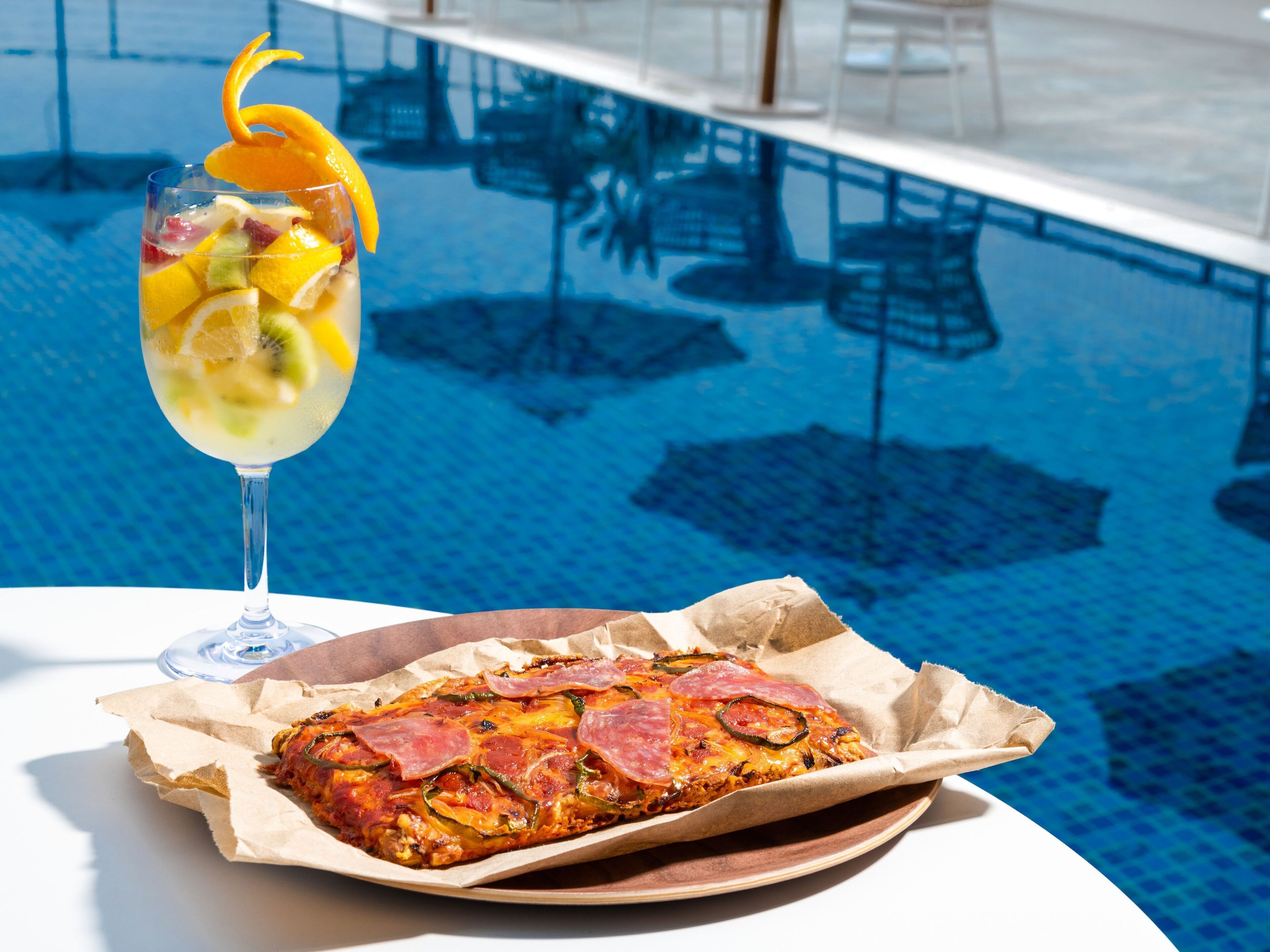 Una mesa junto a la piscina con un vaso de cóctel de frutas y una porción de pizza en un plato de papel.