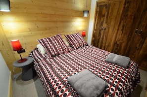 Un dormitorio con cabecera de madera, cama con colcha estampada, dos almohadas, una lámpara roja sobre una mesita de noche y una toalla gris sobre la cama.