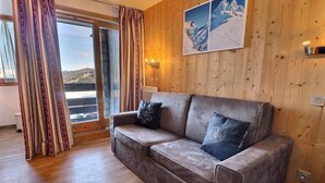 Ett mysigt rum med trägolv, en brun lädersoffa med två kuddar, ett stort fönster med gardiner och en väggmonterad lampa.