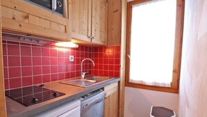 Uma cozinha compacta com armários de madeira, painel de azulejos vermelhos, pia de aço inoxidável e janela com cortinas brancas.