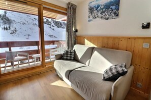 A cozy room with a sofa, two pillows, a wooden floor, a large window with a view of a snowy landscape, and a wall-mounted picture.
