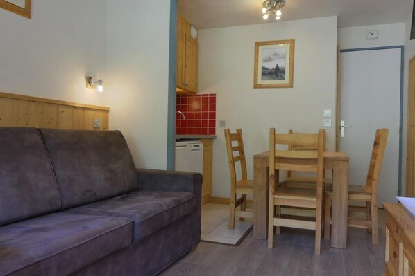 A compact living space with a sofa, wooden dining table, and chairs, a small kitchen area with a red backsplash, and a framed picture on the wall.