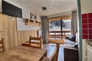 Ein gemütliches Zimmer mit Esstisch aus Holz, Stühlen, einer Couch und einem großen Fenster mit Blick auf eine verschneite Landschaft.