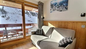 A cozy room with a sofa, two pillows, a wooden floor, a large window with a view of a snowy landscape, and a wall-mounted picture. - 10m from ski slopes, 5th floor, balcony, ski locker, 20m², Méribel-Mottaret (Méribel - Les Allues)