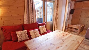 A wooden room with a red sofa, a wooden table, and a window with curtains. - 10m from ski slopes, 3rd floor, balcony, ski locker, 21m², Méribel-Mottaret (Méribel - Les Allues)