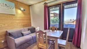A room with a wooden wall, a gray sofa, a wooden dining table with chairs, and a window with curtains. - 10m from ski slopes, 1st floor, balcony, ski locker, 20m², Méribel-Mottaret (Méribel - Les Allues)