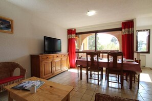 A living room with a television on a wooden cabinet, a dining table with chairs, and a window with red curtains. - 150m from resort center, 2nd floor, view mountain, balcony, parking, tv, 44m², Serre Chevalier (Briançon)