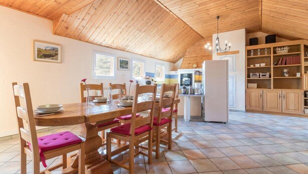 A dining area with a wooden table and chairs, a kitchen with a refrigerator, and a bookshelf.