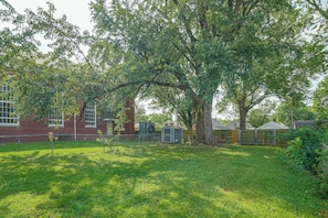 Een omheinde achtertuin met een grote boom, een bakstenen gebouw en een tuinhuis.