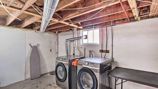 A laundry room with two stacked front-loading washing machines, a metal table, and exposed wooden beams. - Near Churchill Downs: Pet Friendly, Fenced Yard! (Louisville)