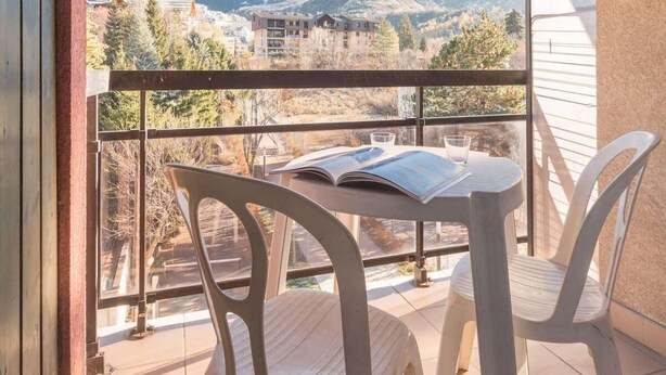 A balcony with a table and chairs, offering a view of a mountain and buildings in the distance.
