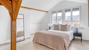 Een slaapkamer met een groot bed, een raam met zonwering, een houten balk in het plafond en een nachtkastje met een lamp.