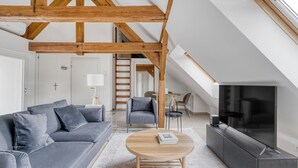 Een moderne loft-woonkamer met zichtbare houten balken, een grijze bank, een salontafel en een televisie.
