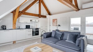 Een moderne keuken met witte kasten, een inbouwoven en een magnetron. In de woonkamer staat een grijze bank met kussens, een ronde houten salontafel en een witte vloerkleed. De ruimte heeft zichtbare houten balken in het plafond.