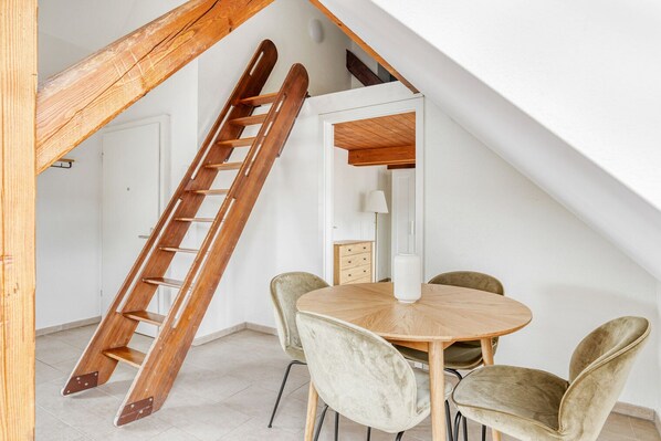 A dining area with a wooden table and chairs, a white lamp, and a wooden ladder against a white wall.