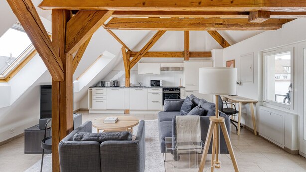 Ruime loft met hoge plafonds, zichtbare houten balken, een moderne keuken, een gezellige zithoek met een bank en fauteuils, en een groot raam dat natuurlijk licht biedt.