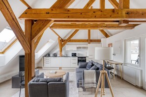 Ein geräumiger Loft mit hohen Decken, freiliegenden Holzbalken, einer modernen Küche, einer gemütlichen Sitzecke mit Sofa und Sesseln sowie einem großen Fenster, das für natürliches Licht sorgt.