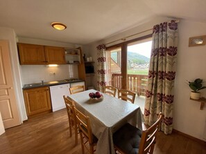 A dining area with a table set for four, a kitchen in the background, and a view of a mountainous landscape through a window.