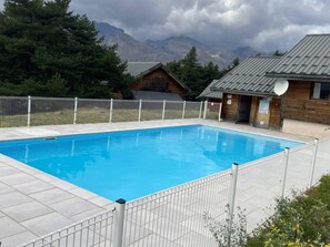 A rectangular swimming pool with a white fence surrounding it, set against a backdrop of mountains and trees.