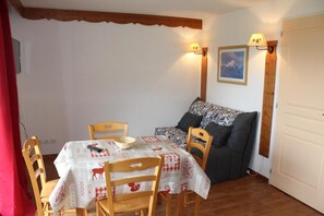 A dining area with a table covered in a checkered tablecloth, wooden chairs, and a sofa with patterned cushions. Two wall-mounted lamps provide lighting.