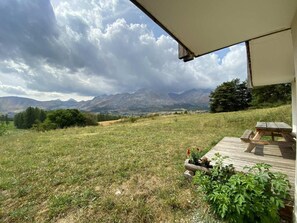 A grassy outdoor area with a wooden deck and picnic table, overlooking a mountainous landscape.