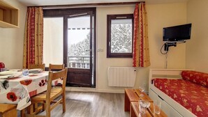 A room with a dining table set for two, a bed with a red patterned bedspread, a television mounted on the wall, and a window with curtains. - 2nd floor, balcony, tv, 24m², Les Menuires (Les Menuires)
