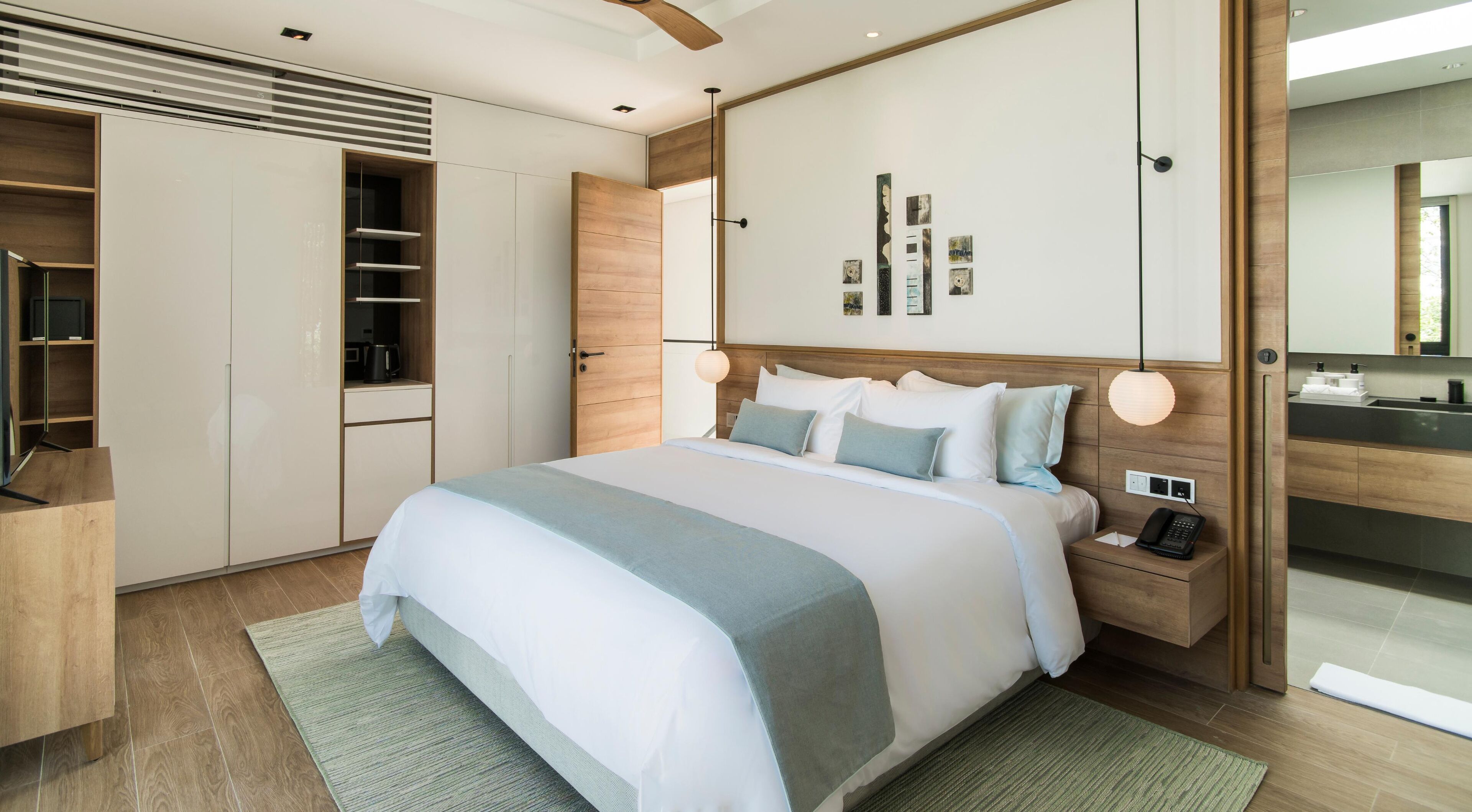 Quarto de hotel moderno com uma cama grande, cabeceira de madeira, armário embutido, mesa de cabeceira com abajur e banheiro visível por uma porta aberta.