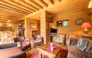 Interior acogedor de una cabaña de troncos, con comedor, sala de estar con sofás y sillones de cuero, televisor y chimenea.