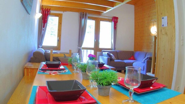 A cozy living room with a wooden ceiling, two windows with curtains, a sofa, a table with red placemats and a laptop, and a potted plant. - 90m from shops, 5th floor, view mountain, balcony, tv, ski locker, 75m², Valmorel (Valmorel)