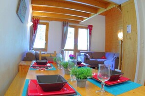 Ein gemütliches Wohnzimmer mit Holzbalkendecke, zwei mit Vorhängen versehenen Fenstern, einem Sofa, einem Tisch mit roten Untersetzern und einem Laptop sowie einer Topfpflanze.