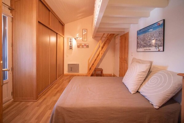 Un dormitorio con un armario de madera, una cama con colcha color beige, un taburete pequeño de madera y un cuadro enmarcado en la pared.