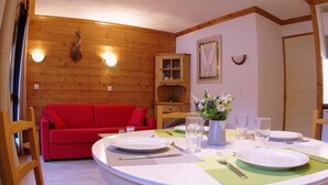 A dining area with a round table set for two, a red sofa, wooden walls, and a cabinet with glass doors. - 240m from ski slopes, 1st floor, view mountain, balcony, ski locker, 37m², Valmorel (Valmorel)