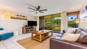 A modern living room with a flat-screen TV, a wooden coffee table, a leather sofa, and a large sliding glass door leading to a garden.