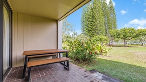 Une terrasse avec un banc en bois, un sol carrelé et une vue sur un espace verdoyant agrémenté d’arbres et de plantes.