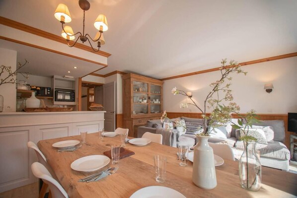 A dining area with a wooden table set for four, a kitchen in the background, and a living area with a sofa and decorative plants.