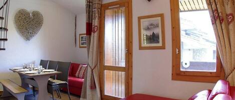 A cozy room with a red sofa, a dining table with chairs, a heart-shaped wall decoration, and a window with curtains.