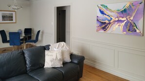 A living room with a black leather sofa, a glass dining table with blue chairs, and a colorful abstract painting on the wall. - French Country Style Main Floor  in the heart of Lethbridge (Lethbridge)