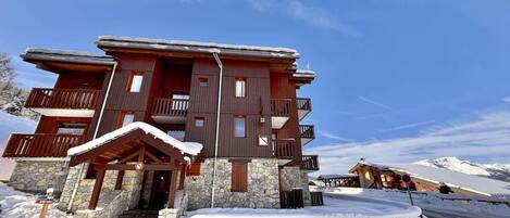 Un bâtiment en bois à plusieurs étages, doté de balcons, d’une fondation en pierre et d’une entrée recouverte de neige.