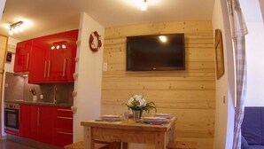 A compact kitchen with red cabinets, a wooden wall-mounted TV, and a dining table with a vase of flowers. - 5m from ski slopes, 1st floor, view valley, balcony, tv, 24m², Valmorel (Valmorel)
