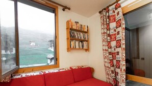 A cozy room with a red sofa, a bookshelf, and a view of a building through a window. - 1st floor, view mountain, tv, 24m², Valmorel (Valmorel)