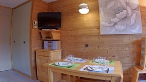 Un interior de madera con una mesa de comedor lista para cuatro comensales, un televisor en un estante y un cuadro grande de flores en la pared.