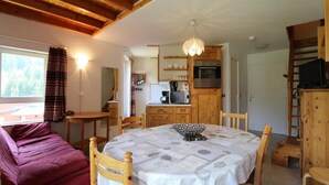 A cozy room with a wooden ceiling, a dining table with a white tablecloth, a purple sofa, and a kitchen area with wooden cabinets and appliances. - 100m from ski slopes, 3rd floor, balcony, tv, ski locker, 35m², Val Cenis (Lanslevillard)