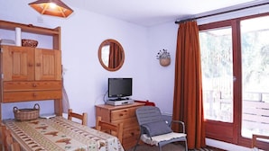 Um quarto com um armário de madeira, uma televisão sobre um suporte de madeira, uma mesa de jantar com toalha de mesa estampada, uma cadeira e uma janela grande com cortinas.