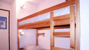 Uma cama beliche com estrutura de madeira e um colchão. Há uma pequena prateleira fixada na estrutura.