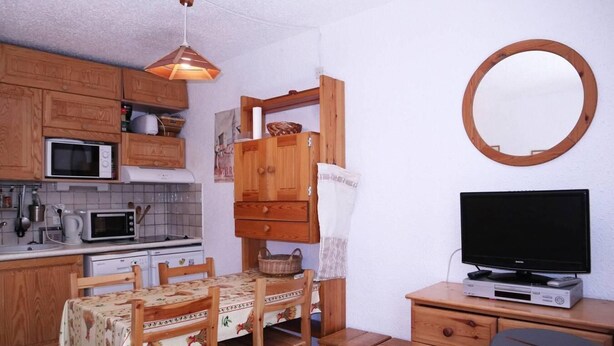 Uma cozinha compacta com armários de madeira, micro-ondas, fogão e uma mesa de jantar com cadeiras. Há uma TV sobre um suporte de madeira e um espelho redondo na parede.