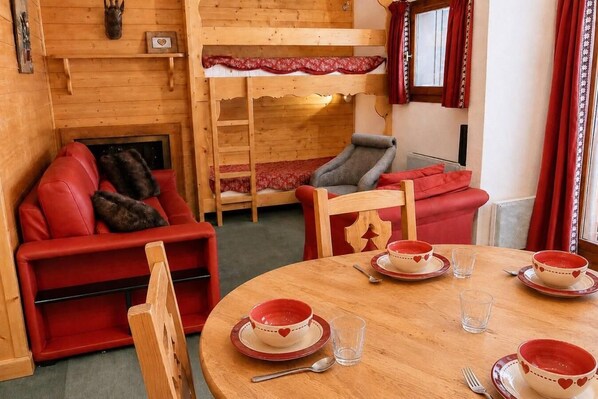 A cozy room with a red sofa, a wooden bunk bed, a dining table with red and white plates, and a window with red curtains.