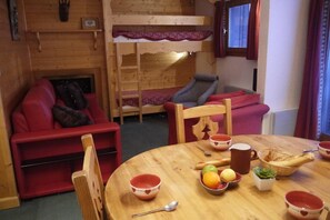 Une pièce chaleureuse avec un lit superposé, un canapé rouge, une table à manger en bois sur laquelle sont disposés des fruits et des bols, et une petite plante en pot.